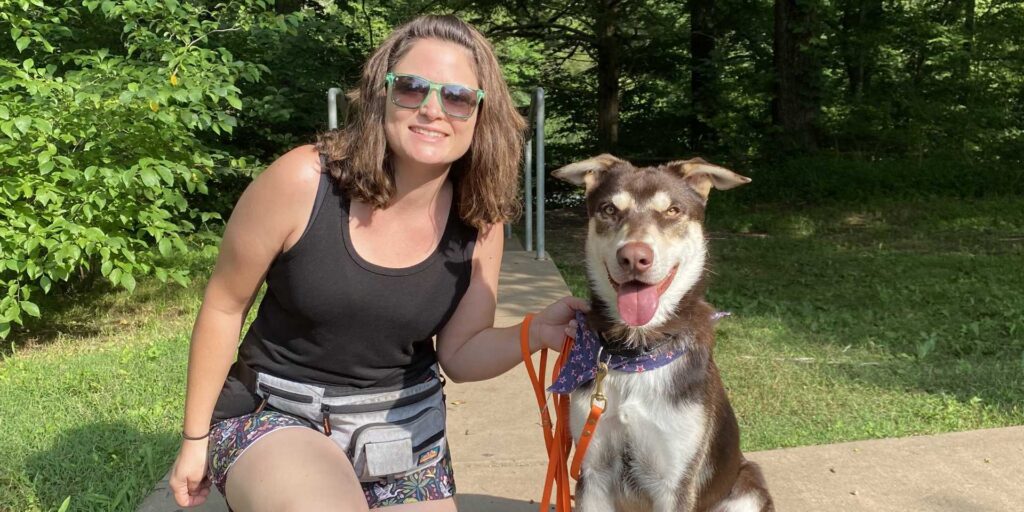 dog trainer gerry-anna in sunglasses sitting outdoors on a paved patio next to a happy brown and white dog on a red leash.